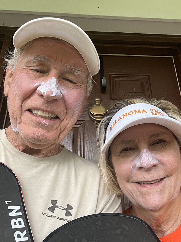 A smiling couple wearing Melanoma Know More visors and sunscreen on their noses.